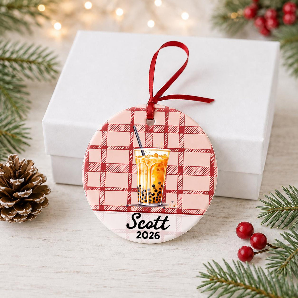 Personalized boba ceramic ornament with name and year on red plaid in front of a white gift box with pinecones and red berries