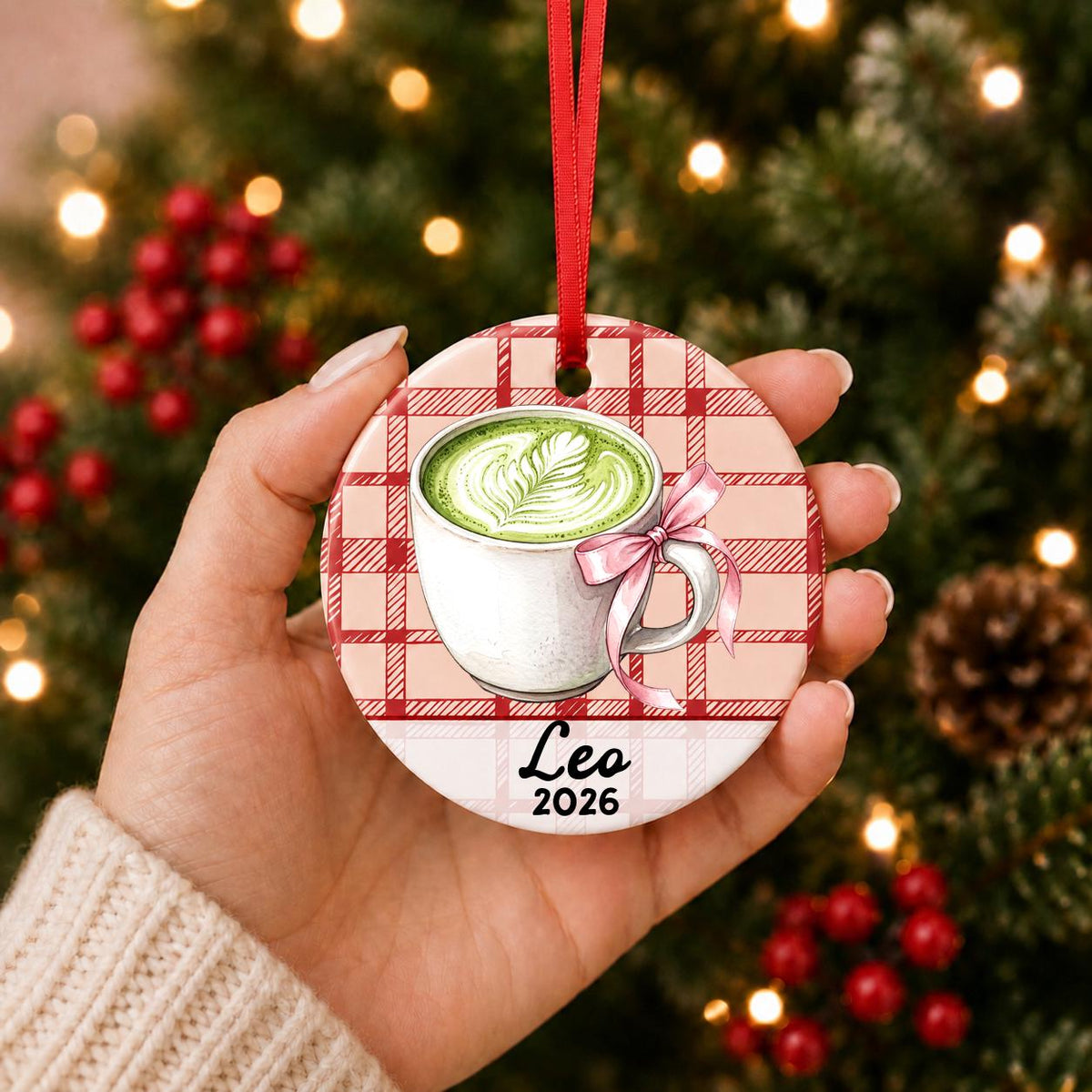 Hand holding the personalized matcha latte ceramic ornament with name and year on red plaid in front of a Christmas tree