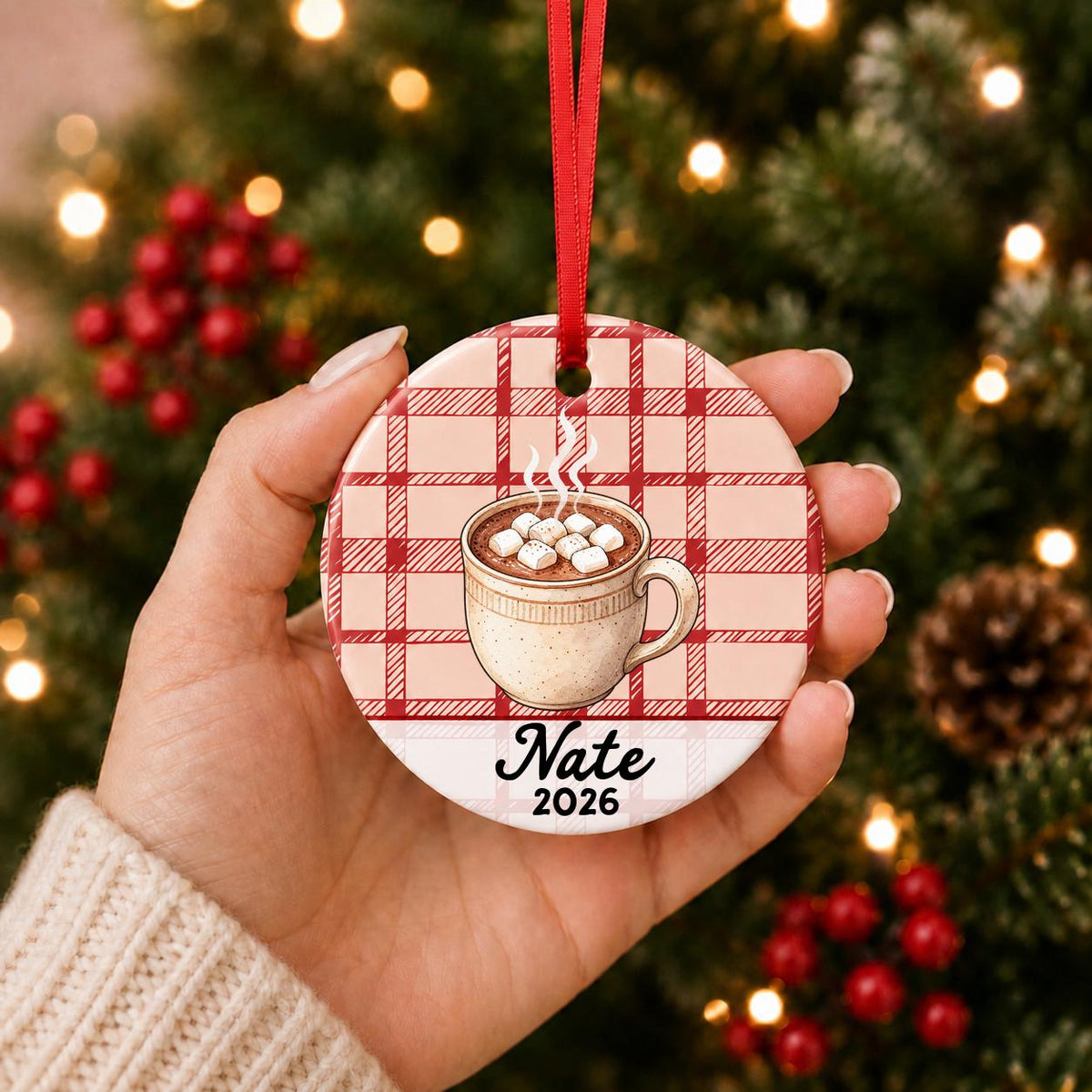 Hand holding the personalized hot chocolate ceramic ornament with name and year on red plaid in front of a Christmas tree