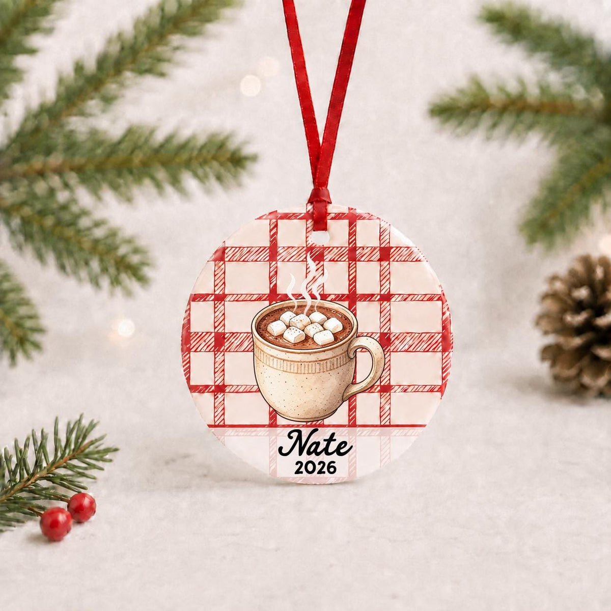 Personalized hot chocolate ceramic ornament with name and year on red plaid with red ribbon on a white surface with pine branches and pinecones