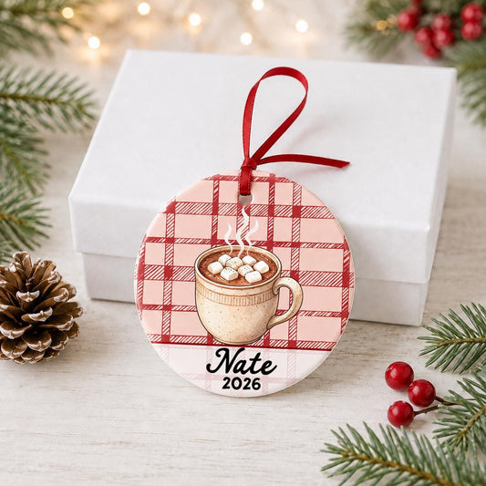 Personalized hot chocolate ceramic ornament with name and year on red plaid in front of a white gift box with pinecones and red berries