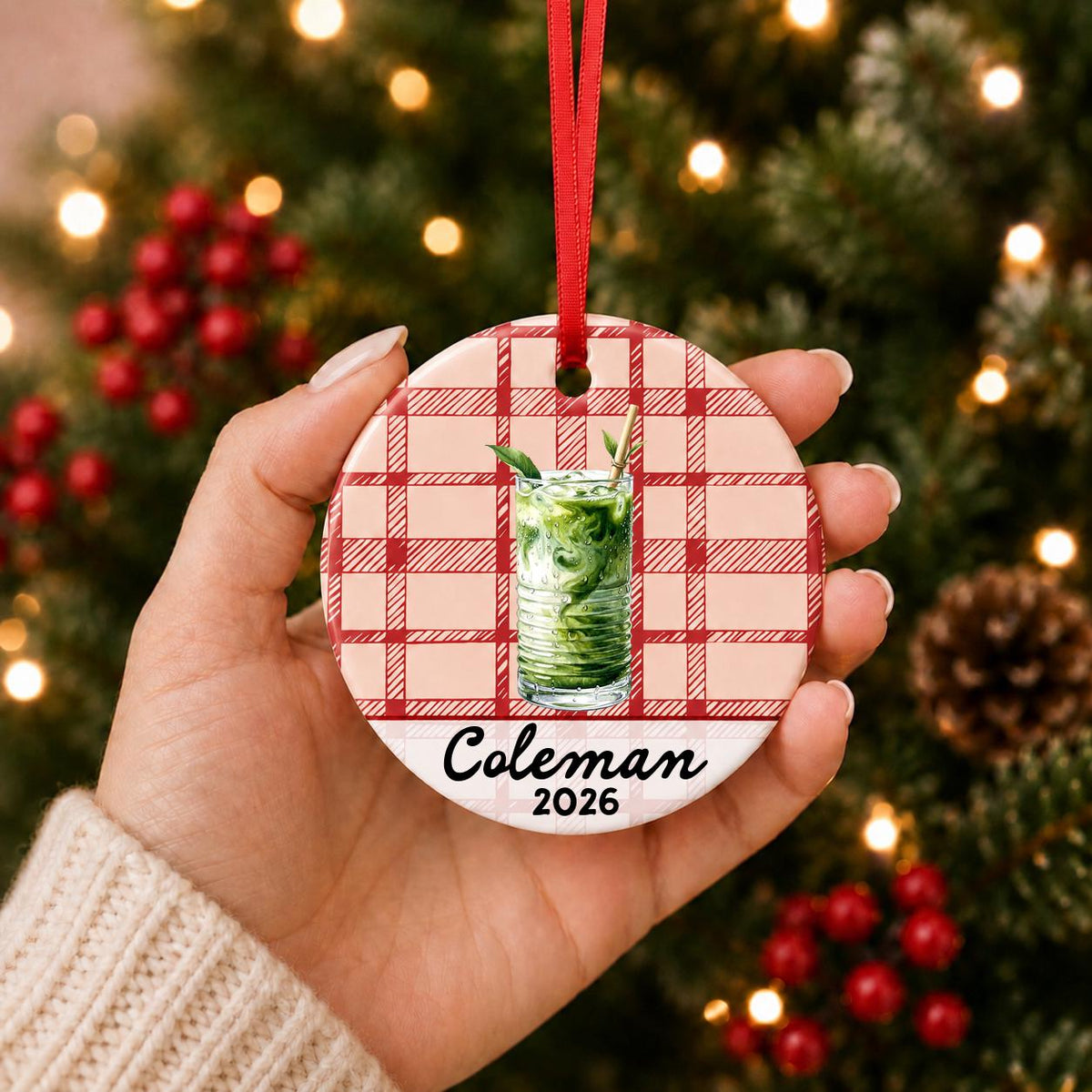 Hand holding the personalized iced matcha ceramic ornament with name and year on red plaid in front of a Christmas tree
