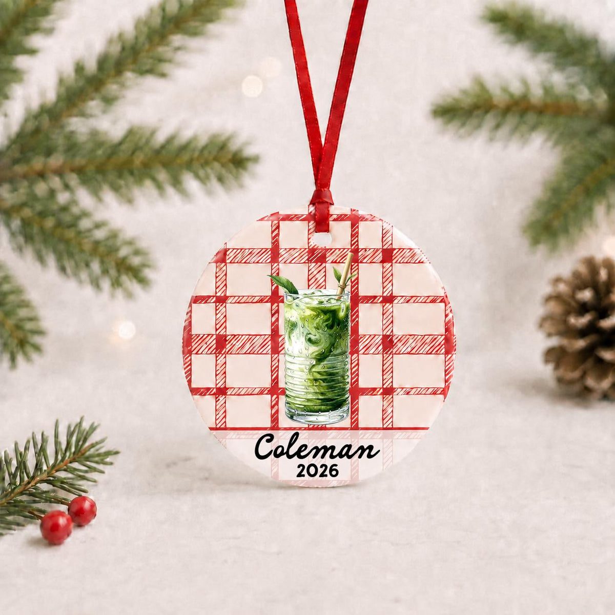 Personalized iced matcha ceramic ornament with name and year on red plaid with red ribbon on a white surface with pine branches and pinecones