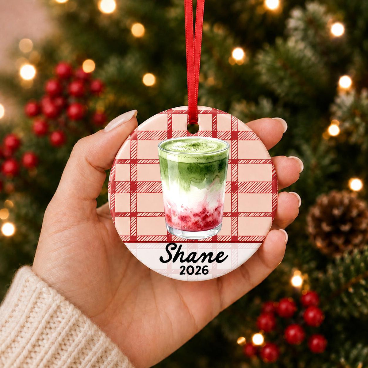 Hand holding the personalized strawberry matcha latte ceramic ornament with name and year on red plaid in front of a Christmas tree