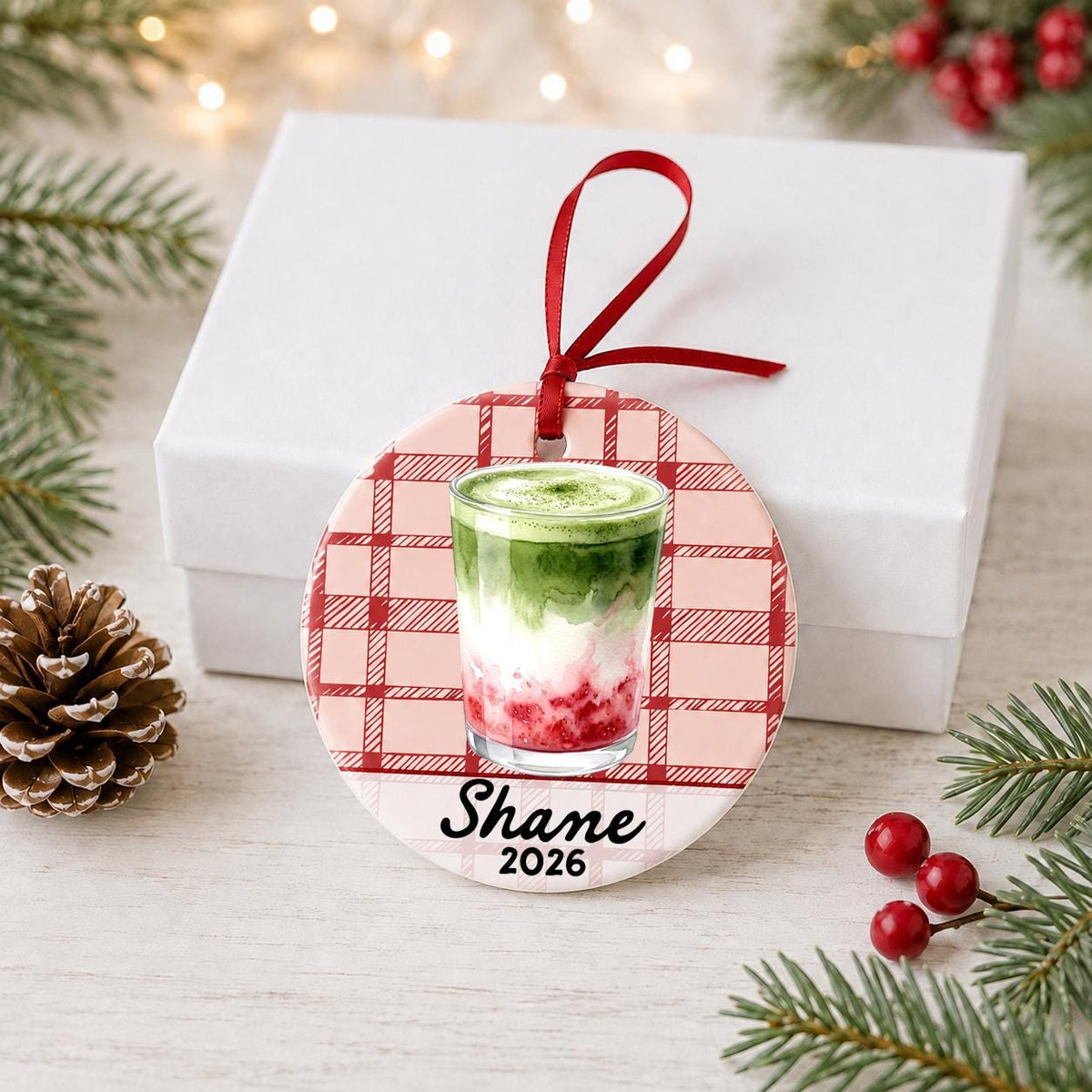 Personalized strawberry matcha latte ceramic ornament with name and year on red plaid in front of a white gift box with pinecones and red berries