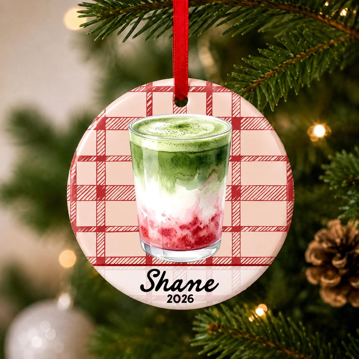 Close-up of the personalized strawberry matcha latte ceramic ornament with name and year on red plaid hanging on a Christmas tree