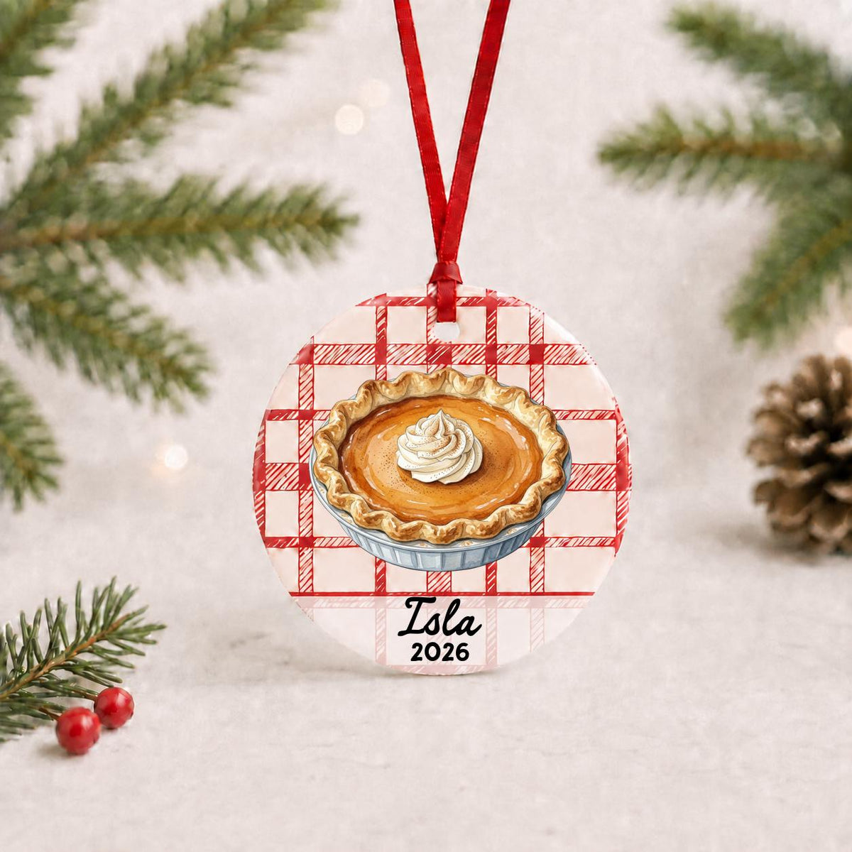 Personalized pumpkin pie ceramic ornament with name and year on red plaid with red ribbon on a white surface with pine branches and pinecones