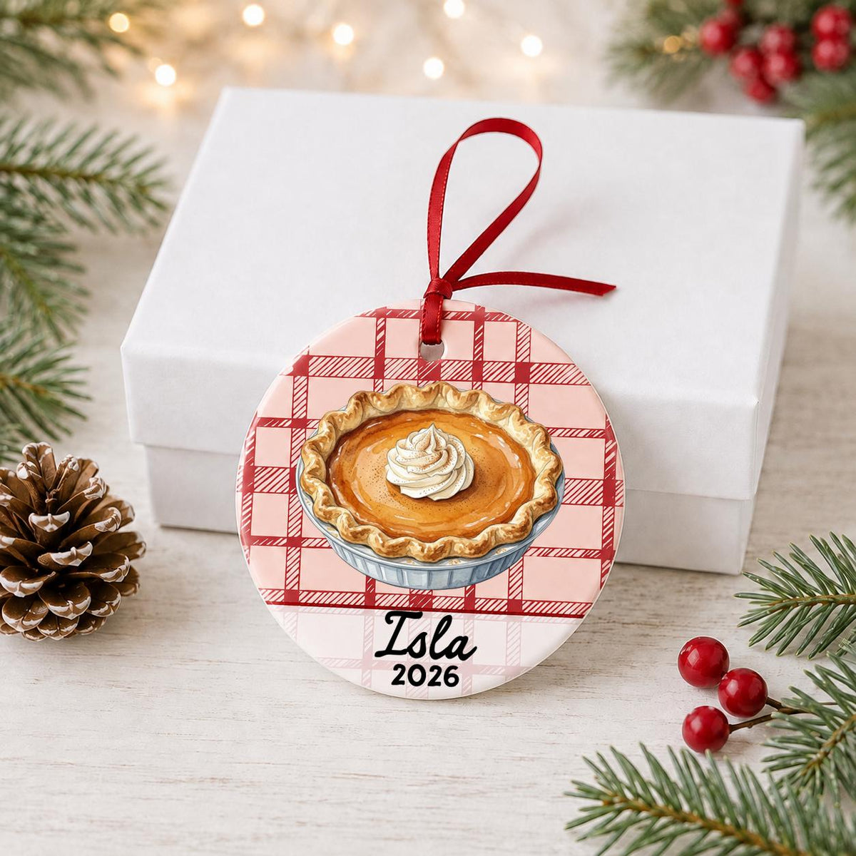 Personalized pumpkin pie ceramic ornament with name and year on red plaid in front of a white gift box with pinecones and red berries