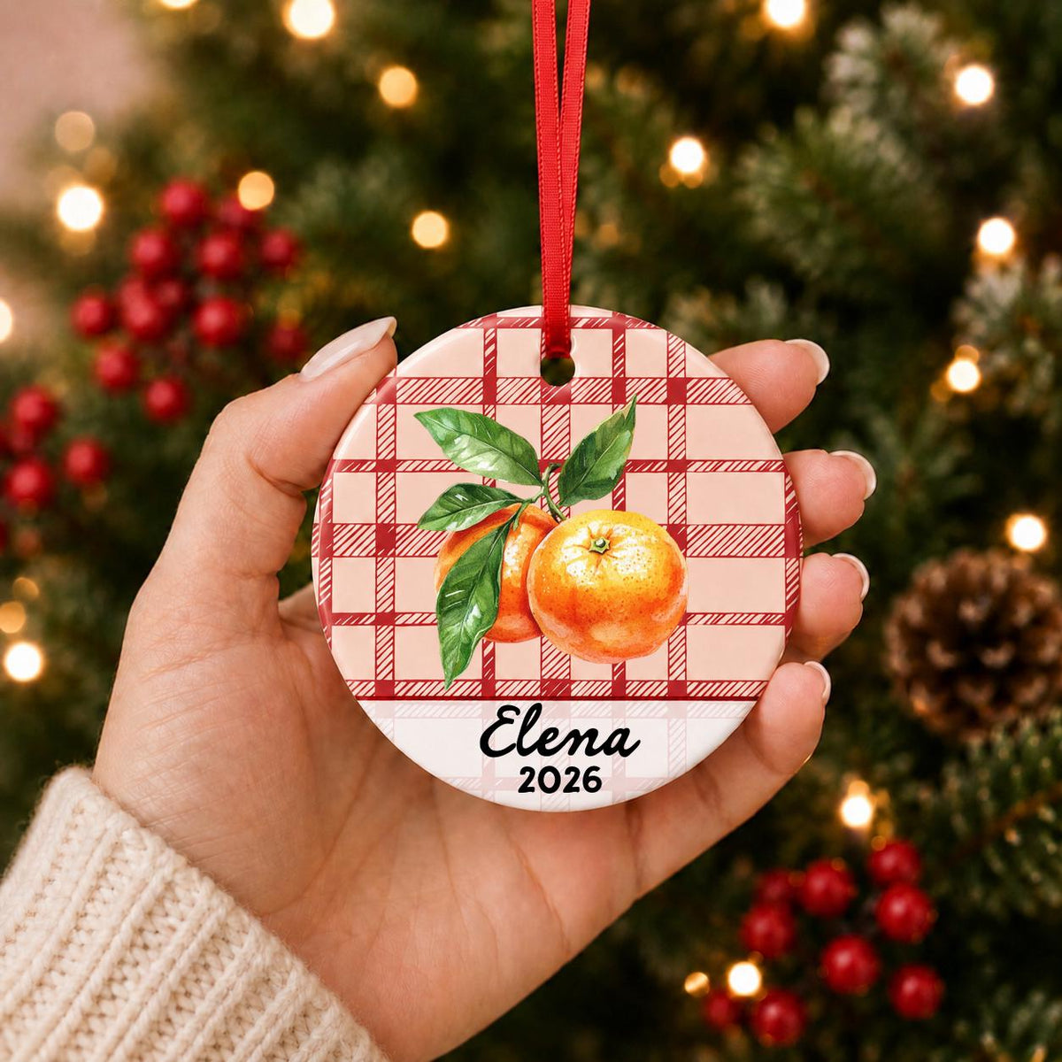 Hand holding the personalized clementine ceramic ornament with name and year on red plaid in front of a Christmas tree