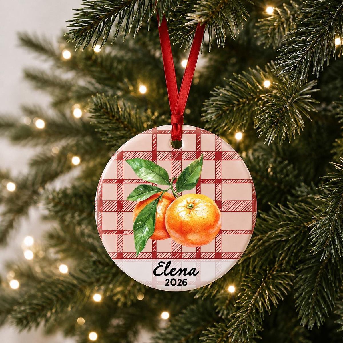 Personalized clementine ceramic ornament with name and year on red plaid hanging on a lit Christmas tree