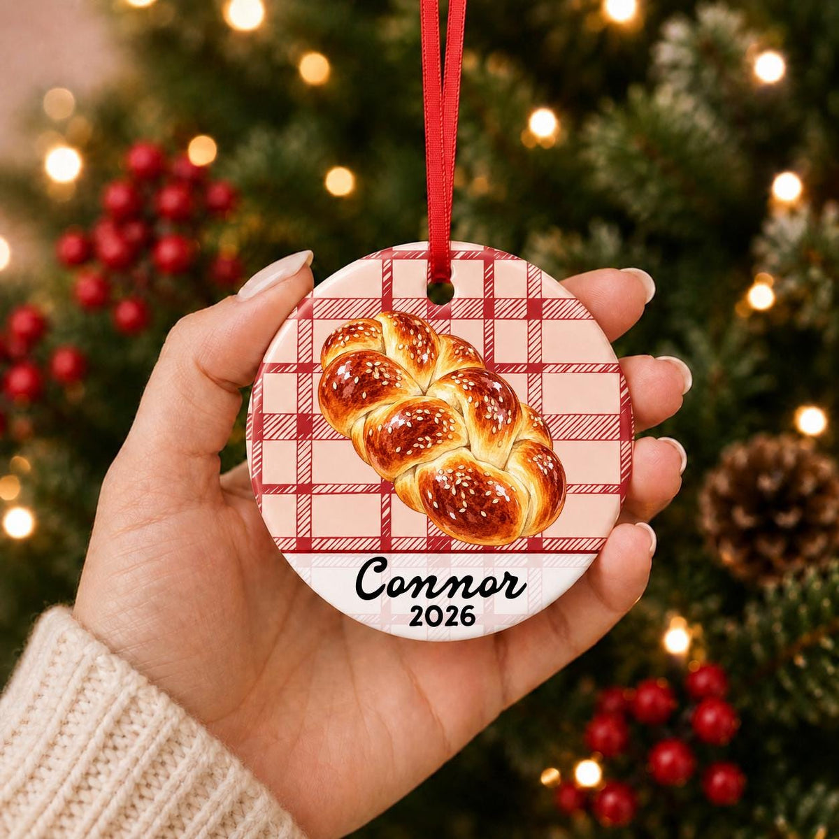 Hand holding the personalized challah bread ceramic ornament with name and year on red plaid in front of a Christmas tree