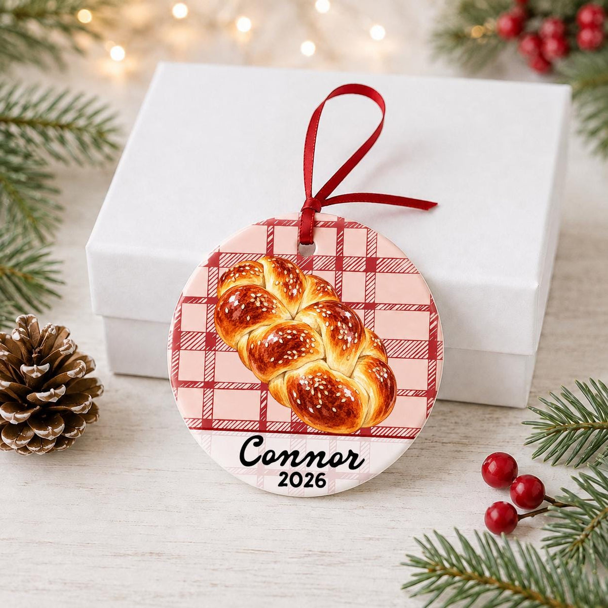 Personalized challah bread ceramic ornament with name and year on red plaid in front of a white gift box with pinecones and red berries