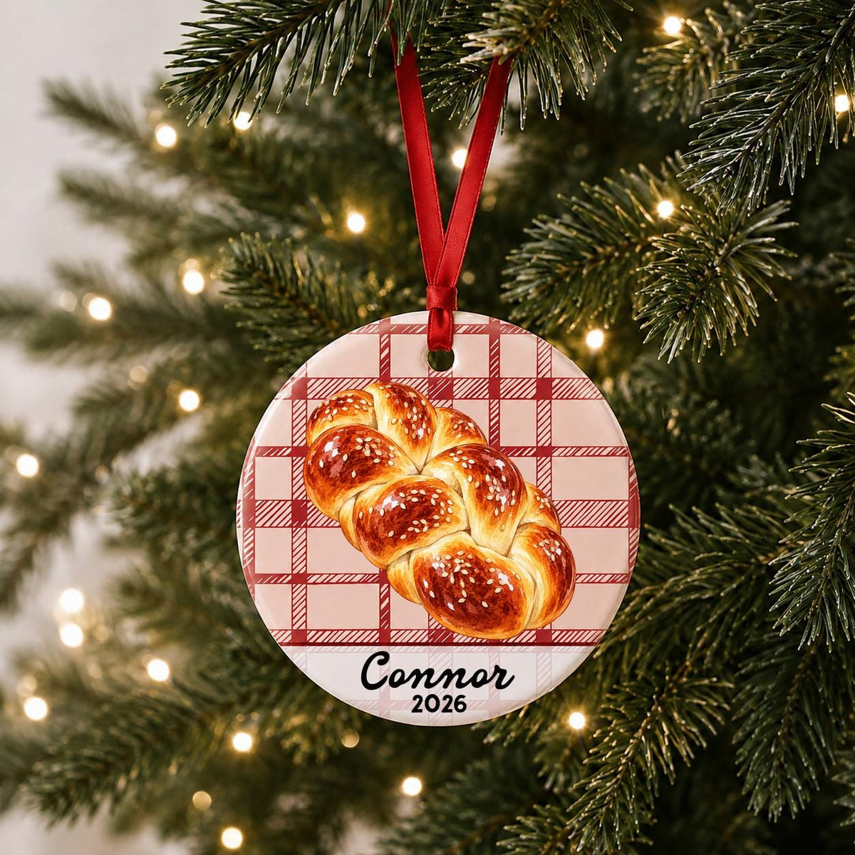 Personalized challah bread ceramic ornament with name and year on red plaid hanging on a lit Christmas tree