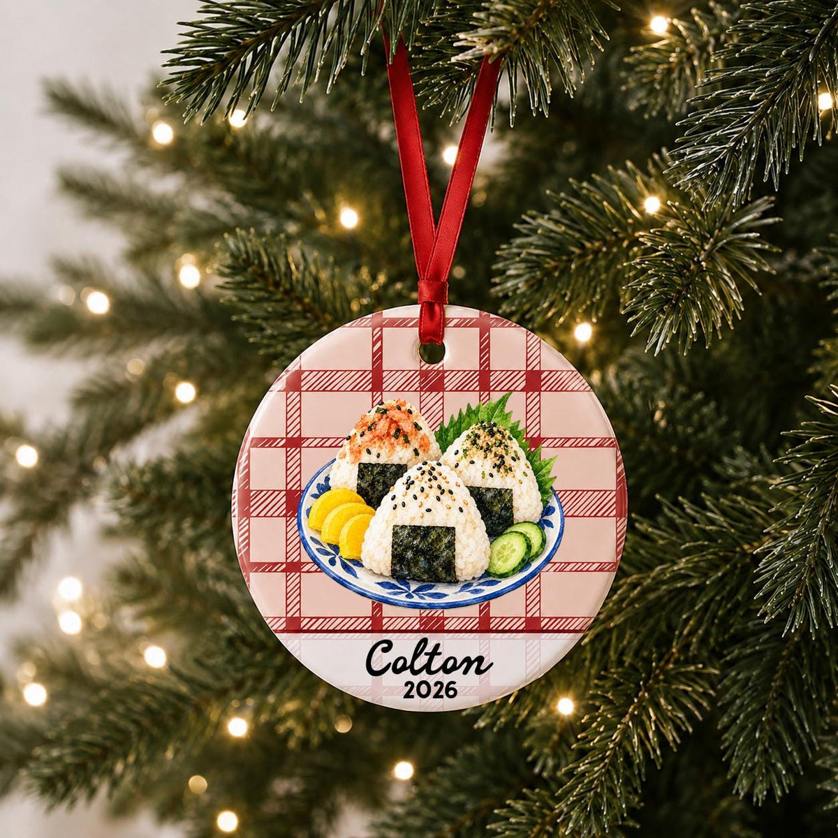 Personalized onigiri ceramic ornament with name and year on red plaid hanging on a lit Christmas tree