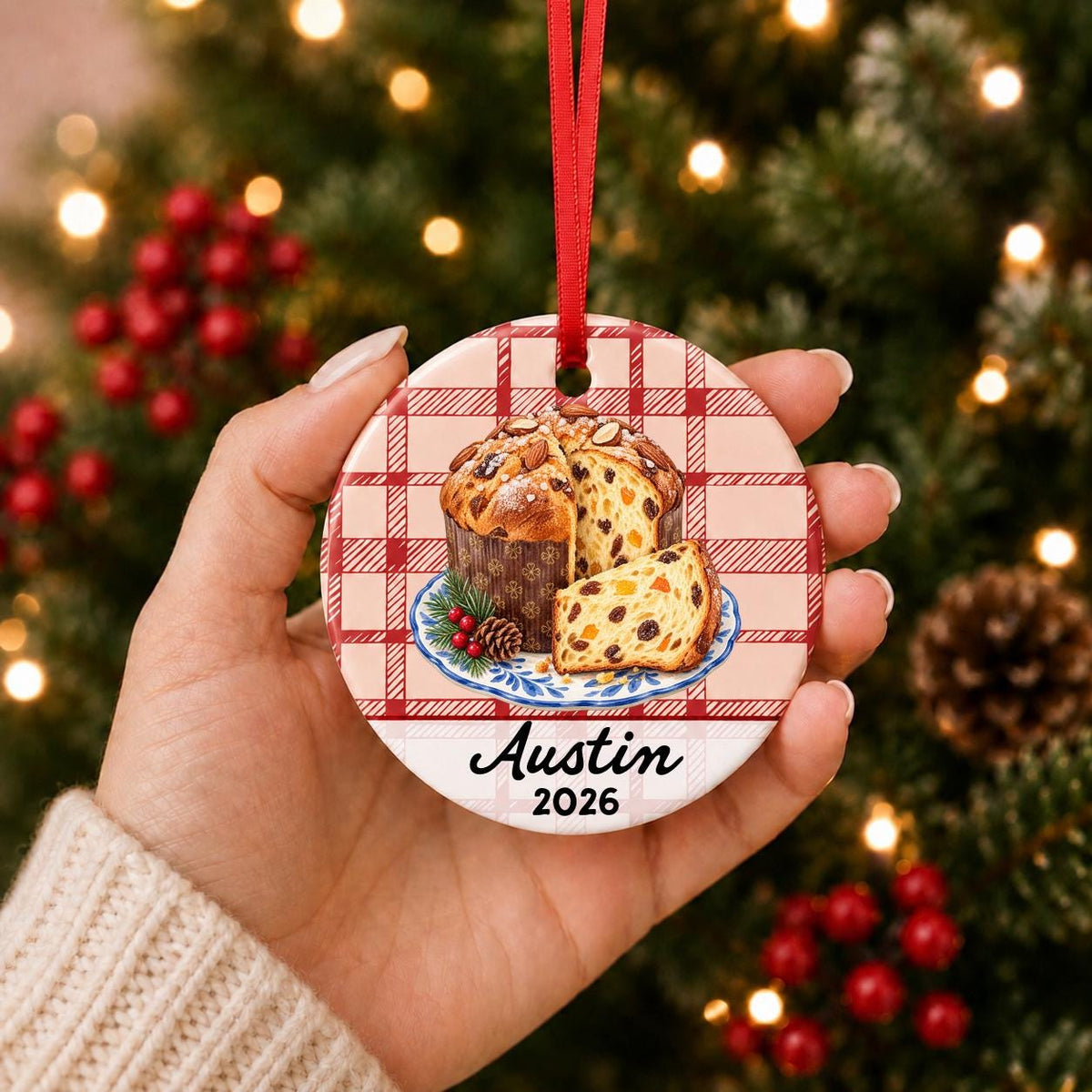 Hand holding the personalized panettone ceramic ornament with name and year on red plaid in front of a Christmas tree