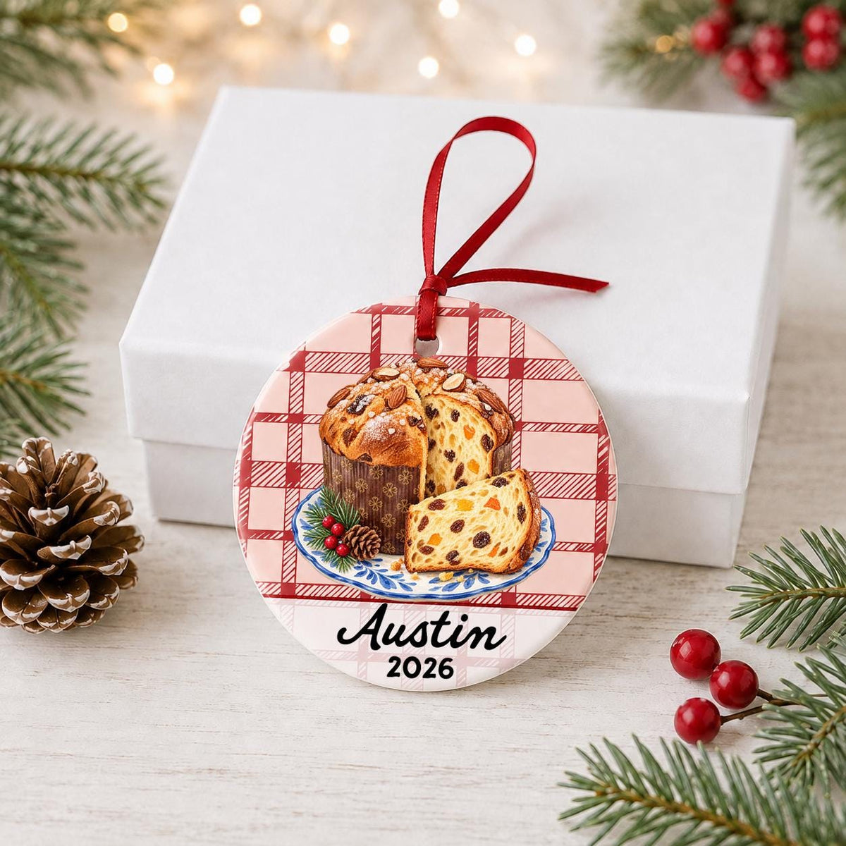 Personalized panettone ceramic ornament with name and year on red plaid in front of a white gift box with pinecones and red berries