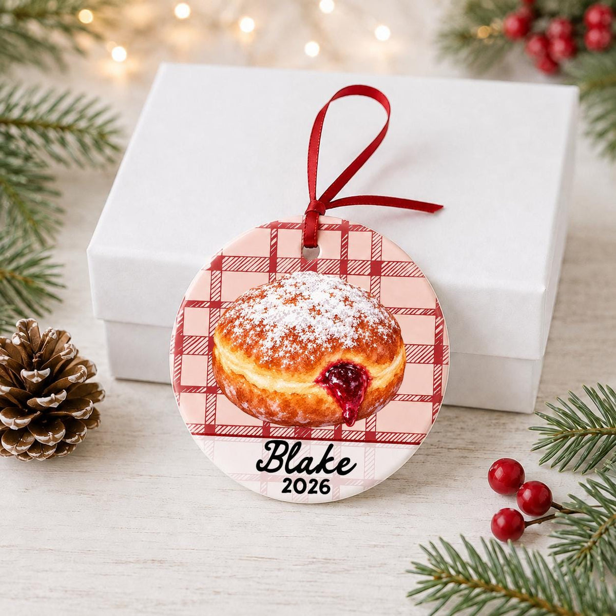 Personalized paczki ceramic ornament with name and year on red plaid in front of a white gift box with pinecones and red berries