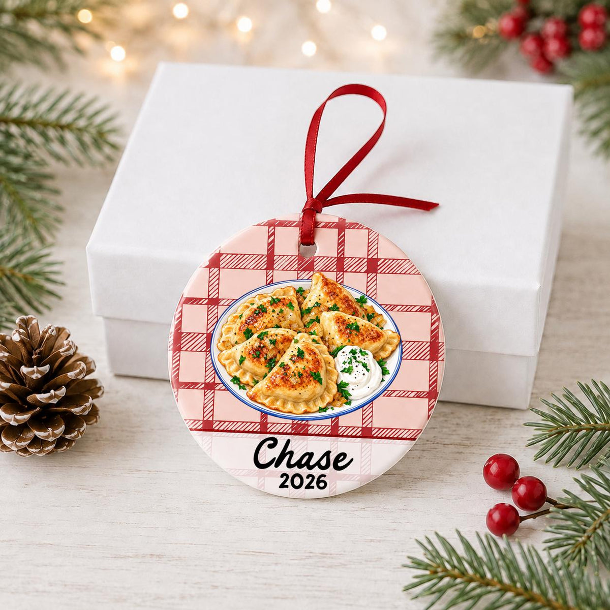 Personalized pierogi ceramic ornament with name and year on red plaid in front of a white gift box with pinecones and red berries