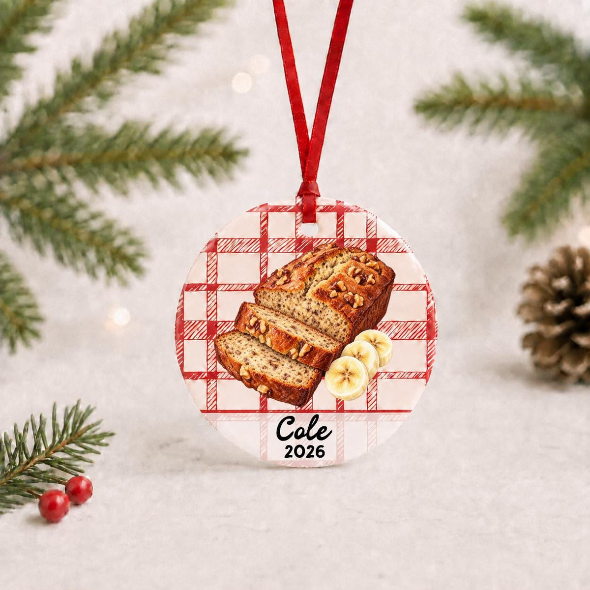 Personalized banana bread ceramic ornament with name and year on red plaid with red ribbon on a white surface with pine branches and pinecones