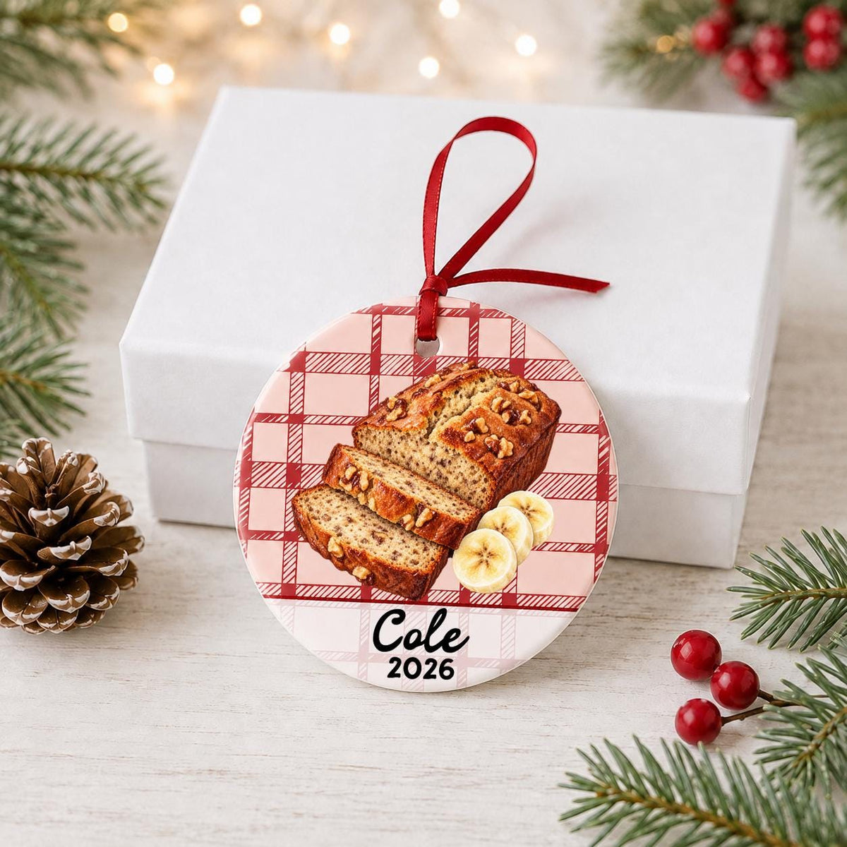 Personalized banana bread ceramic ornament with name and year on red plaid in front of a white gift box with pinecones and red berries