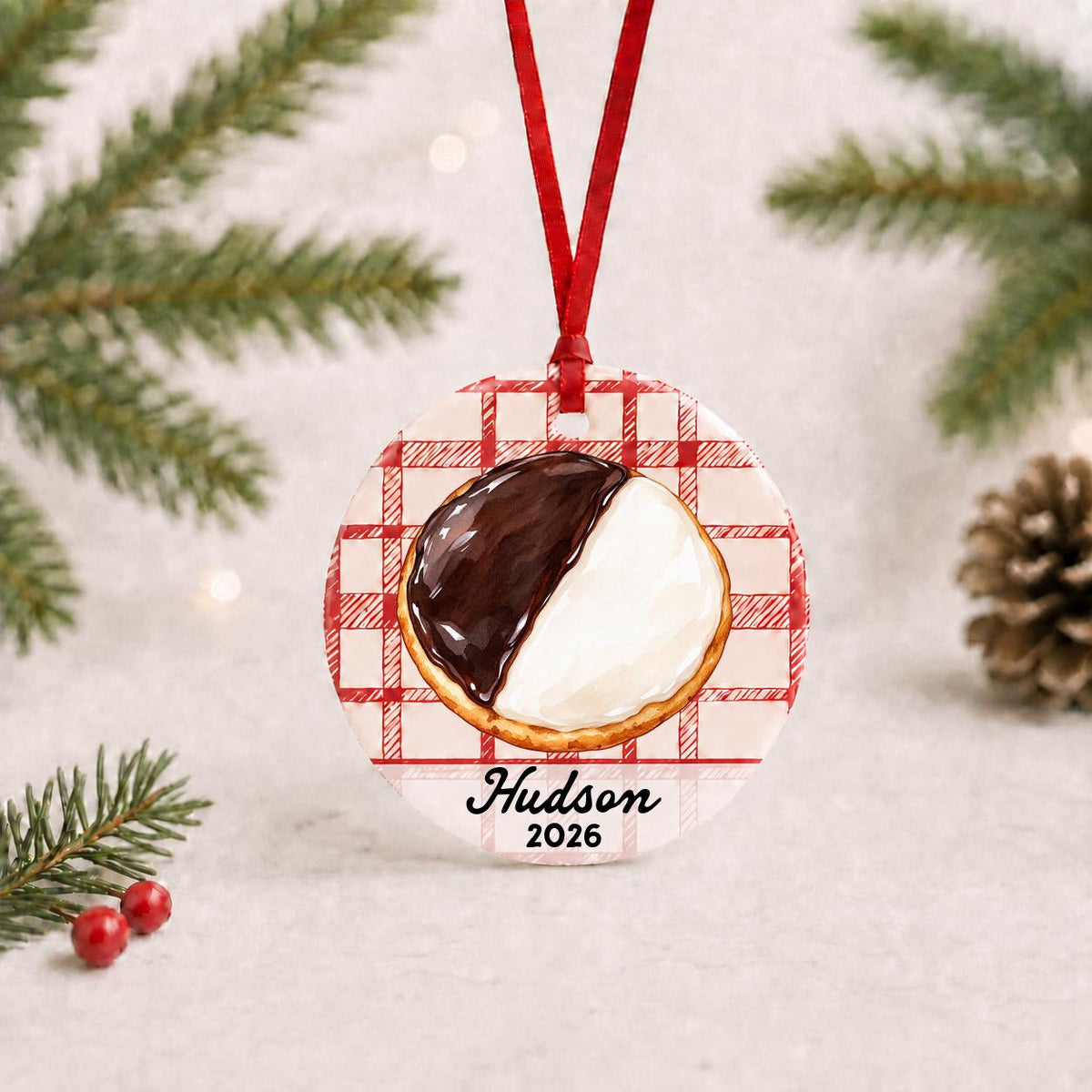 Personalized black and white cookie ceramic ornament with name and year on red plaid with red ribbon on a white surface with pine branches and pinecones