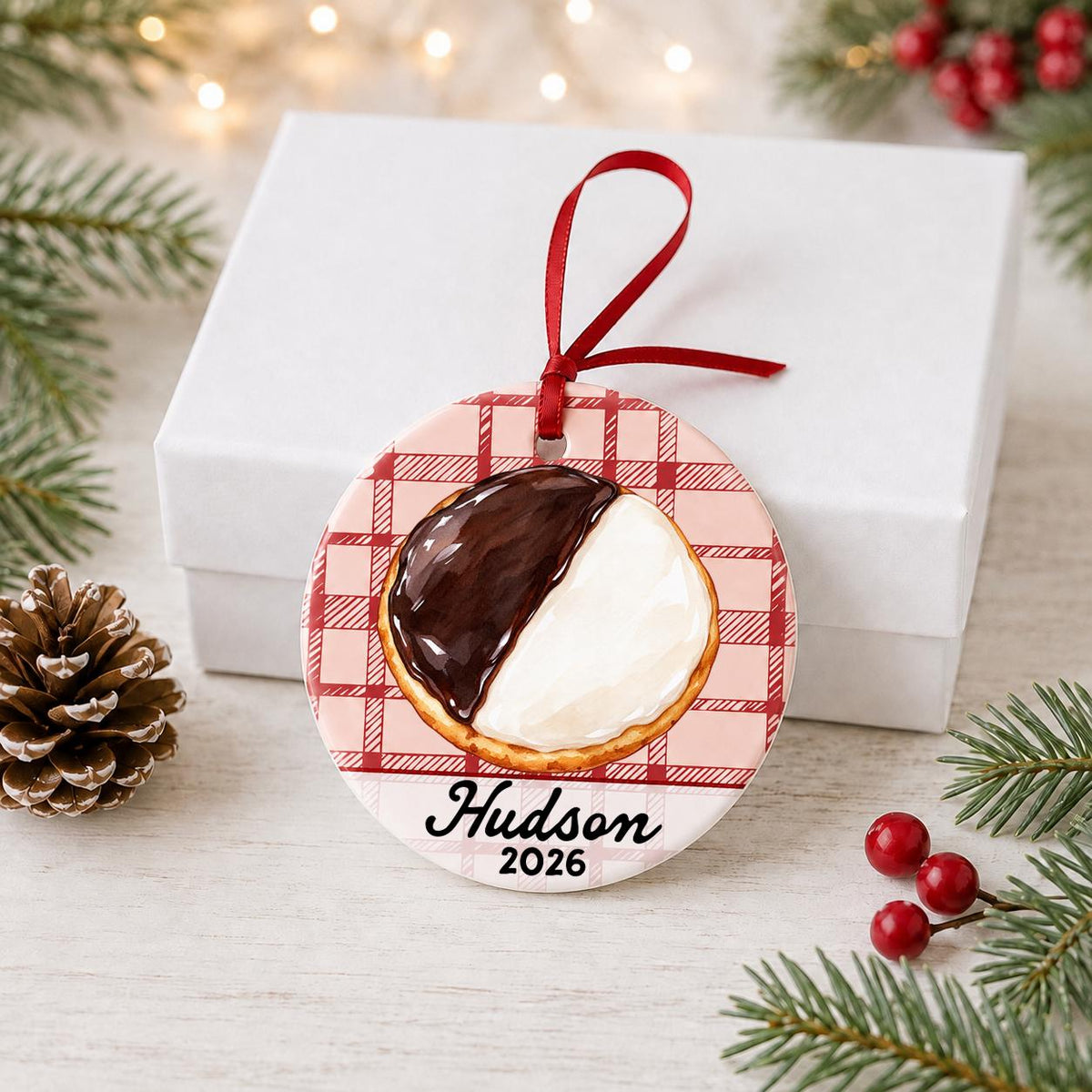 Personalized black and white cookie ceramic ornament with name and year on red plaid in front of a white gift box with pinecones and red berries