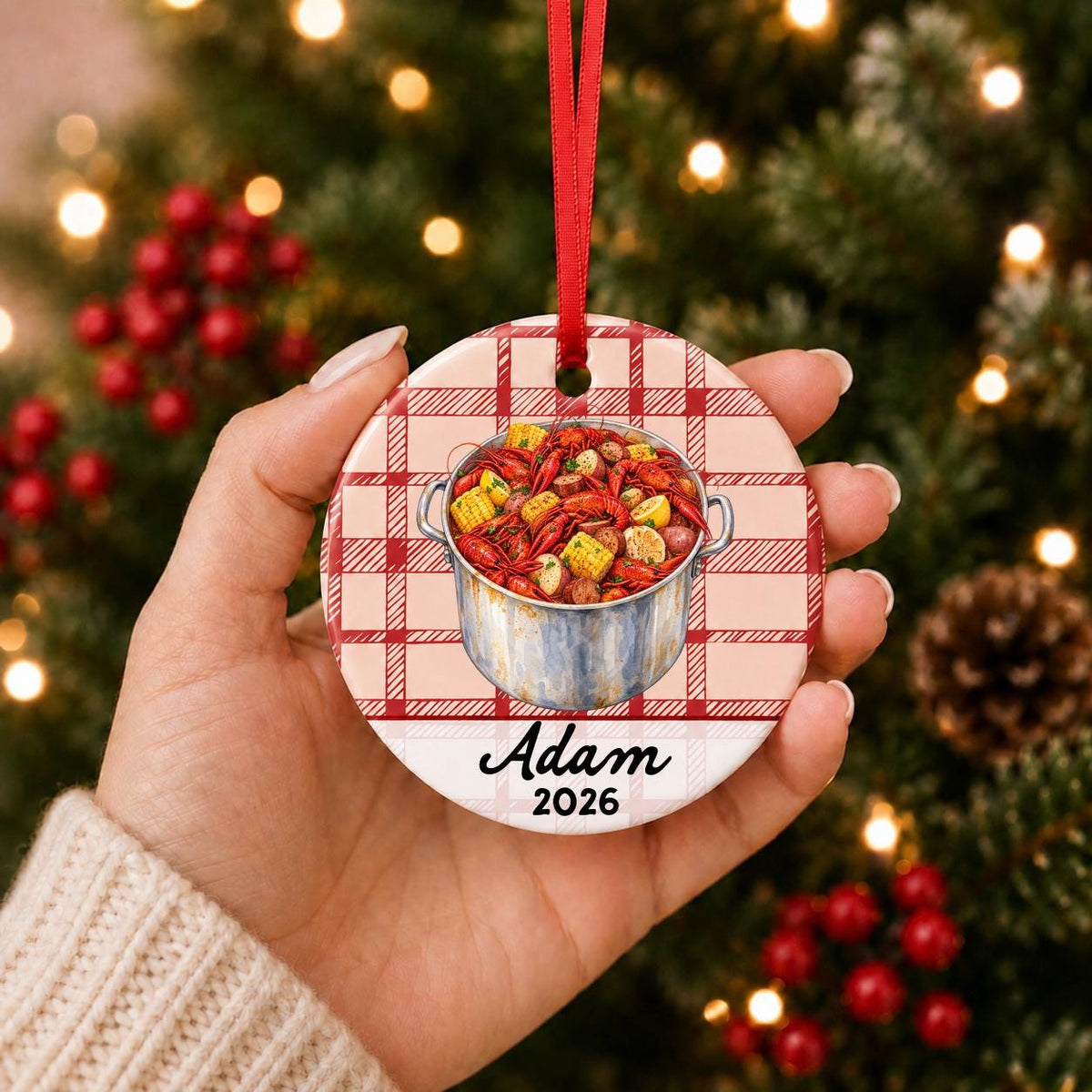 Hand holding the personalized crawfish boil ceramic ornament with name and year on red plaid in front of a Christmas tree