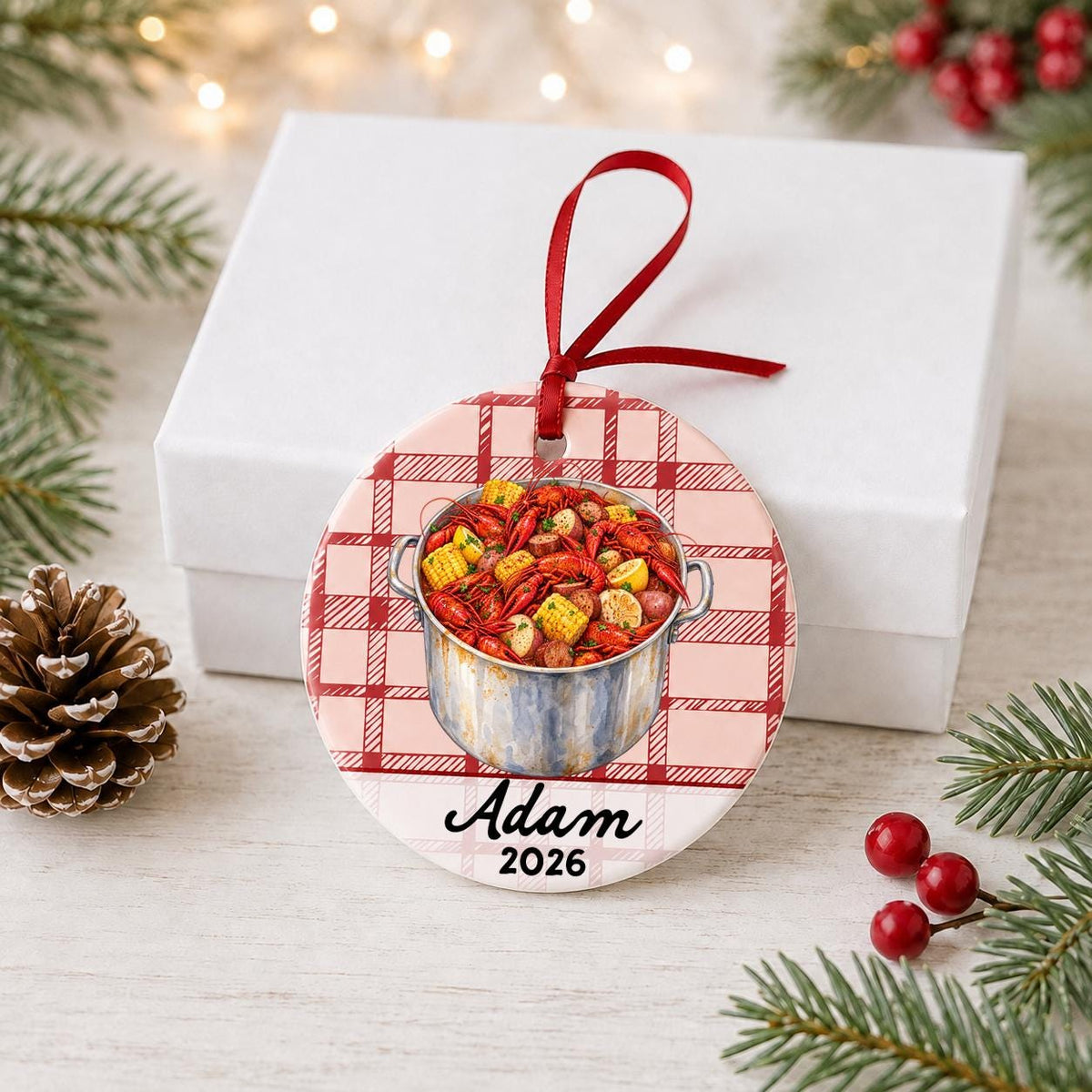 Personalized crawfish boil ceramic ornament with name and year on red plaid in front of a white gift box with pinecones and red berries