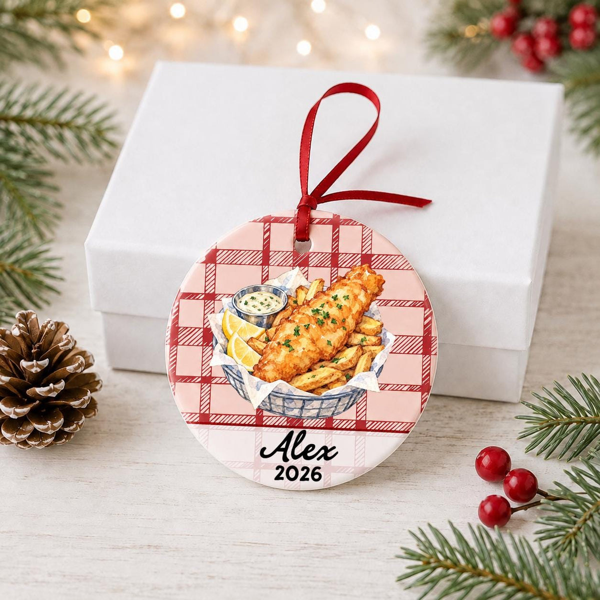Personalized fish and chips ceramic ornament with name and year on red plaid in front of a white gift box with pinecones and red berries