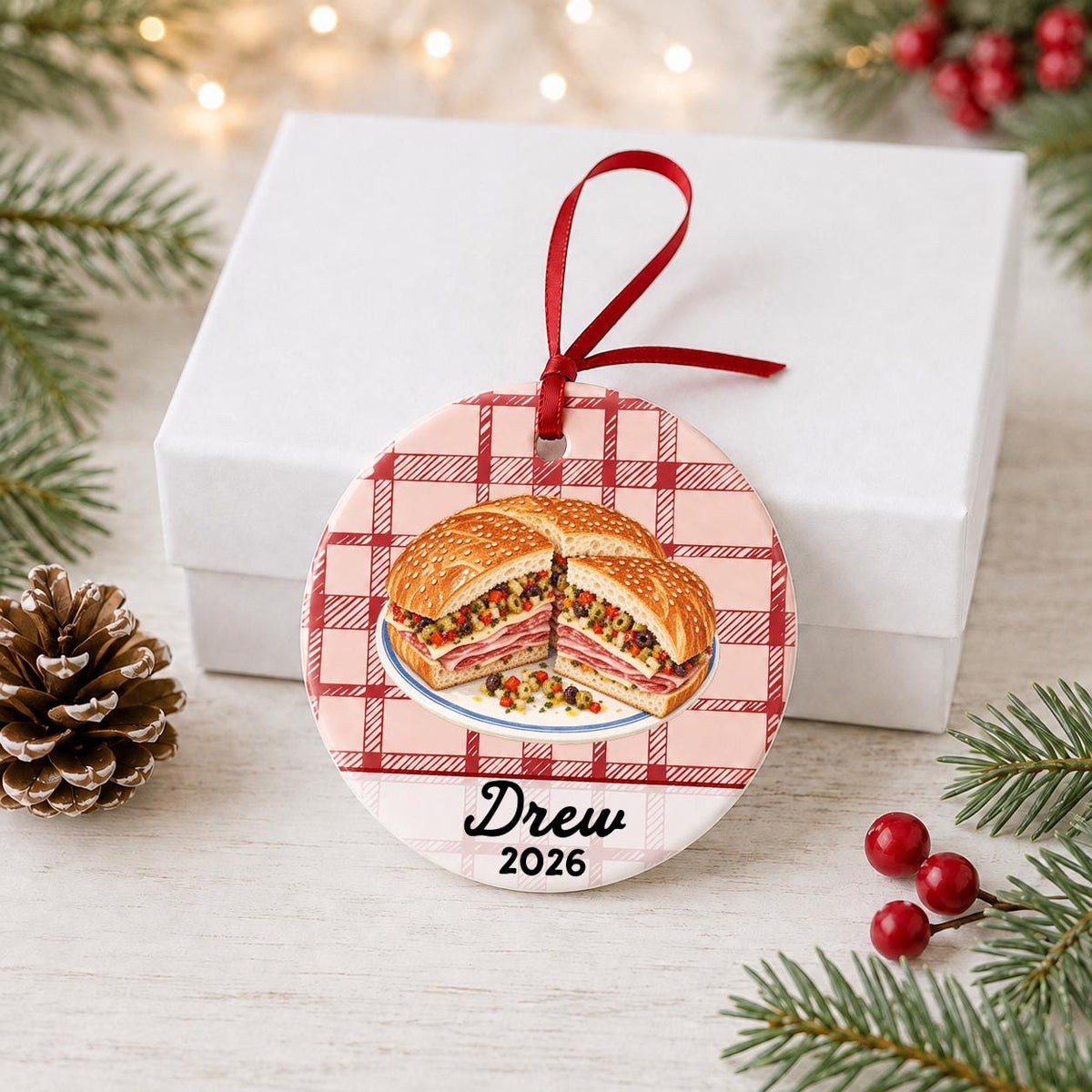 Personalized muffuletta ceramic ornament with name and year on red plaid in front of a white gift box with pinecones and red berries