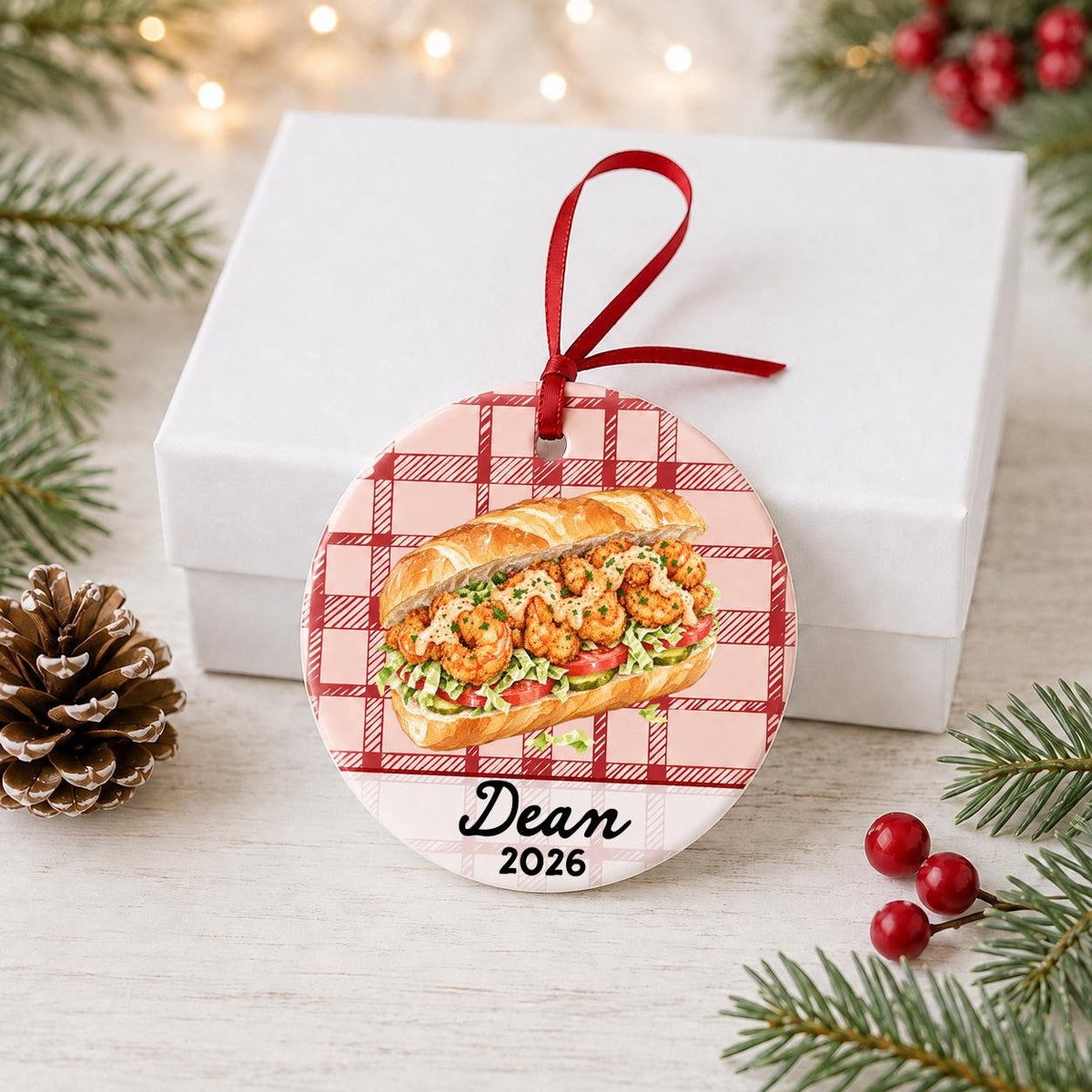 Personalized shrimp po' boy ceramic ornament with name and year on red plaid in front of a white gift box with pinecones and red berries