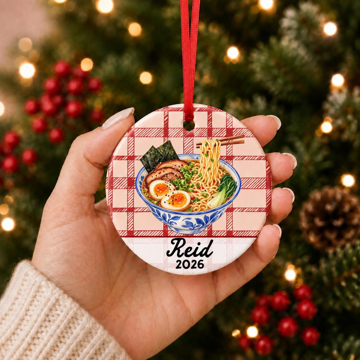 Hand holding the personalized ramen ceramic ornament with name and year on red plaid in front of a Christmas tree