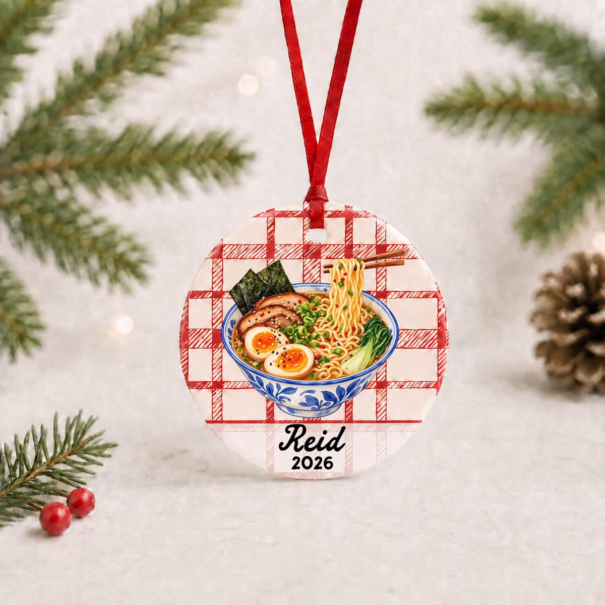 Personalized ramen ceramic ornament with name and year on red plaid with red ribbon on a white surface with pine branches and pinecones