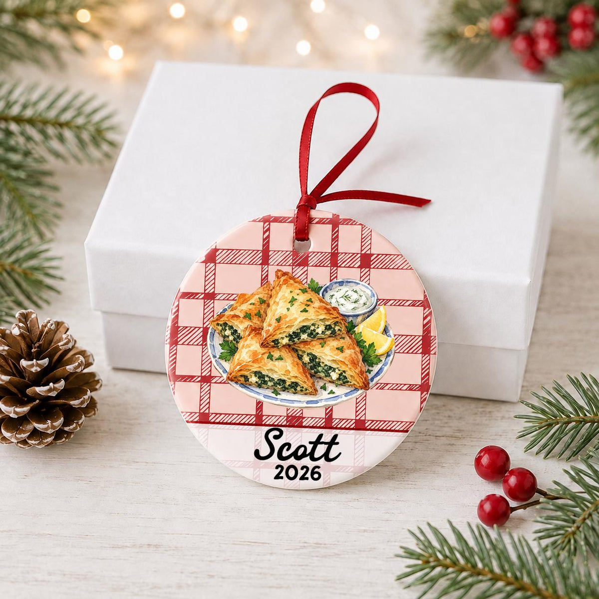 Personalized spanakopita ceramic ornament with name and year on red plaid in front of a white gift box with pinecones and red berries