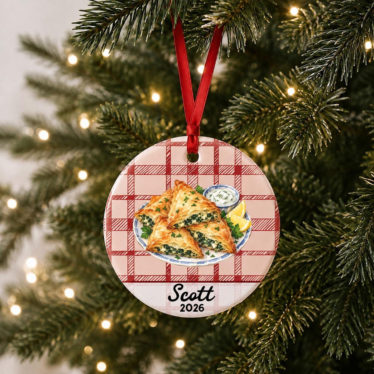 Personalized spanakopita ceramic ornament with name and year on red plaid hanging on a lit Christmas tree