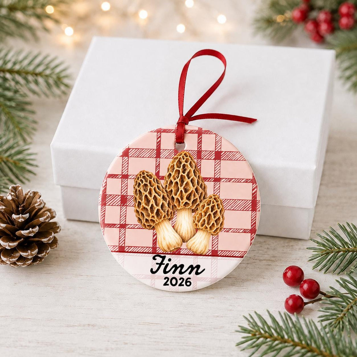 Personalized morel mushrooms ceramic ornament with name and year on red plaid in front of a white gift box with pinecones and red berries
