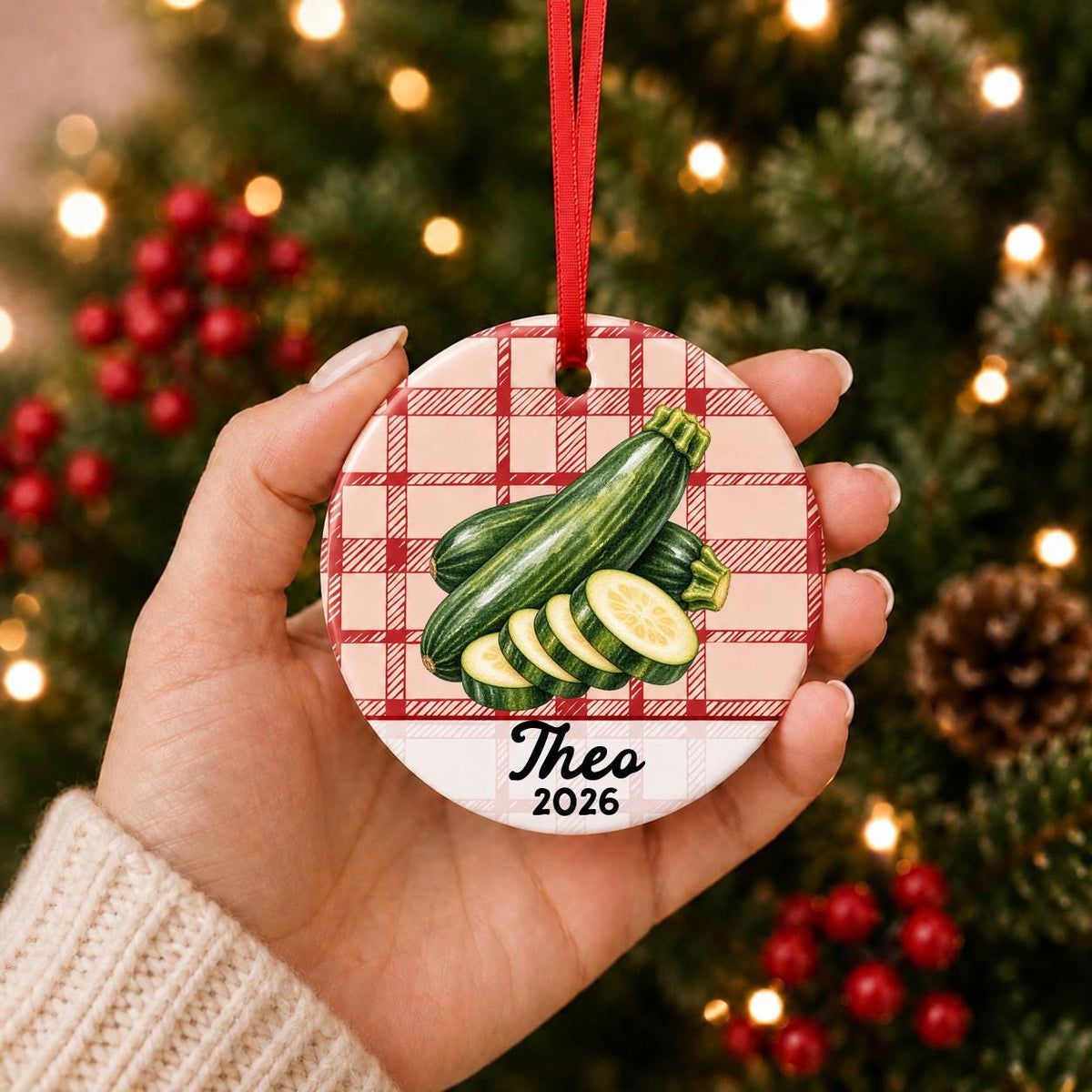 Hand holding the personalized zucchini ceramic ornament with name and year on red plaid in front of a Christmas tree