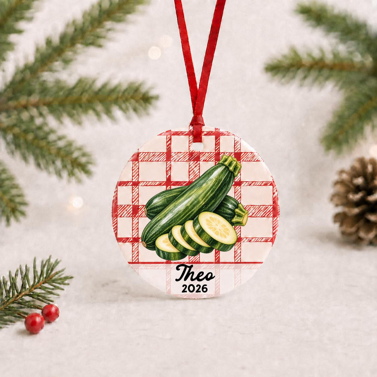 Personalized zucchini ceramic ornament with name and year on red plaid with red ribbon on a white surface with pine branches and pinecones