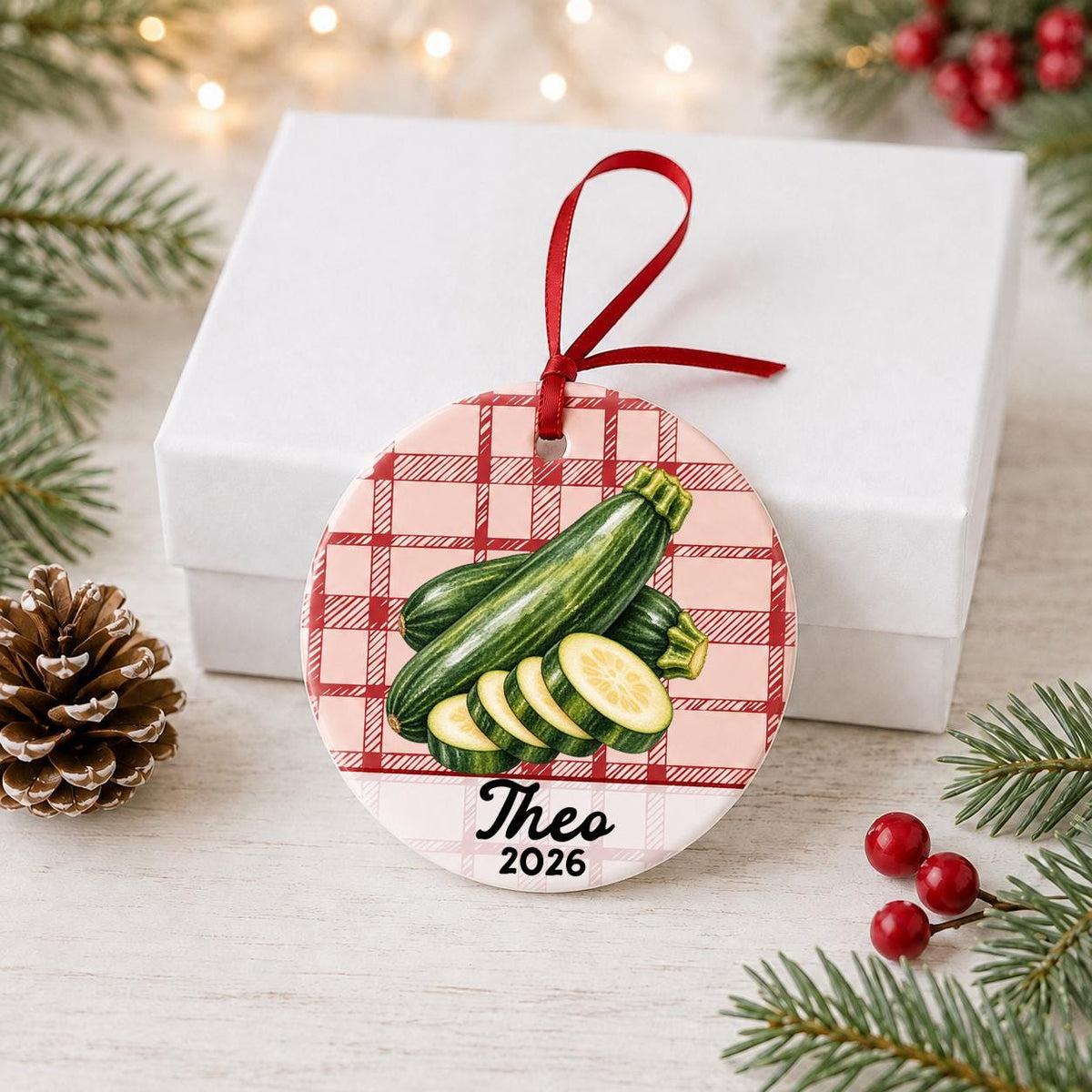 Personalized zucchini ceramic ornament with name and year on red plaid in front of a white gift box with pinecones and red berries