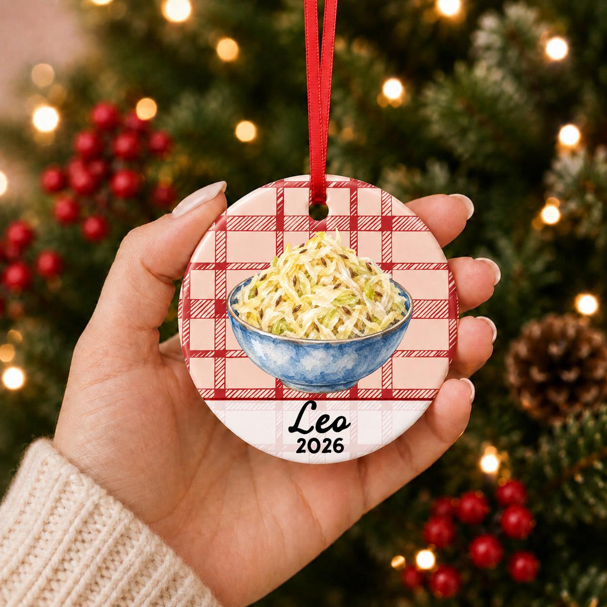 Hand holding the personalized sauerkraut ceramic ornament with name and year on red plaid in front of a Christmas tree