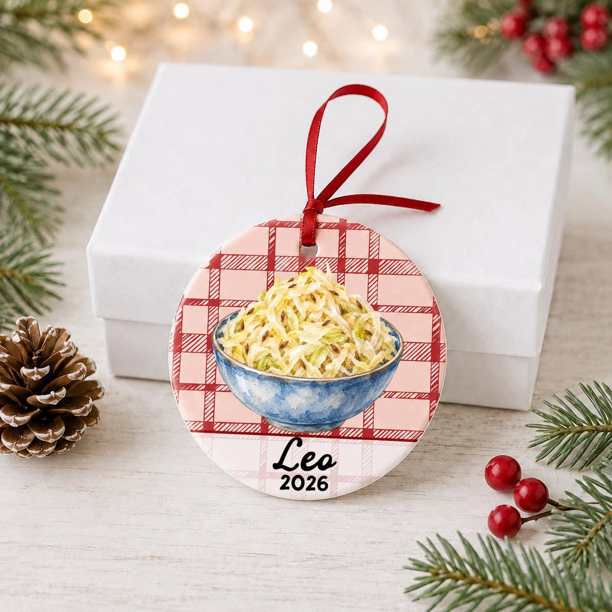 Personalized sauerkraut ceramic ornament with name and year on red plaid in front of a white gift box with pinecones and red berries