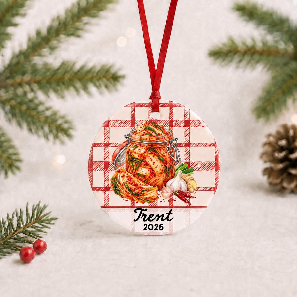 Personalized kimchi ceramic ornament with name and year on red plaid with red ribbon on a white surface with pine branches and pinecones