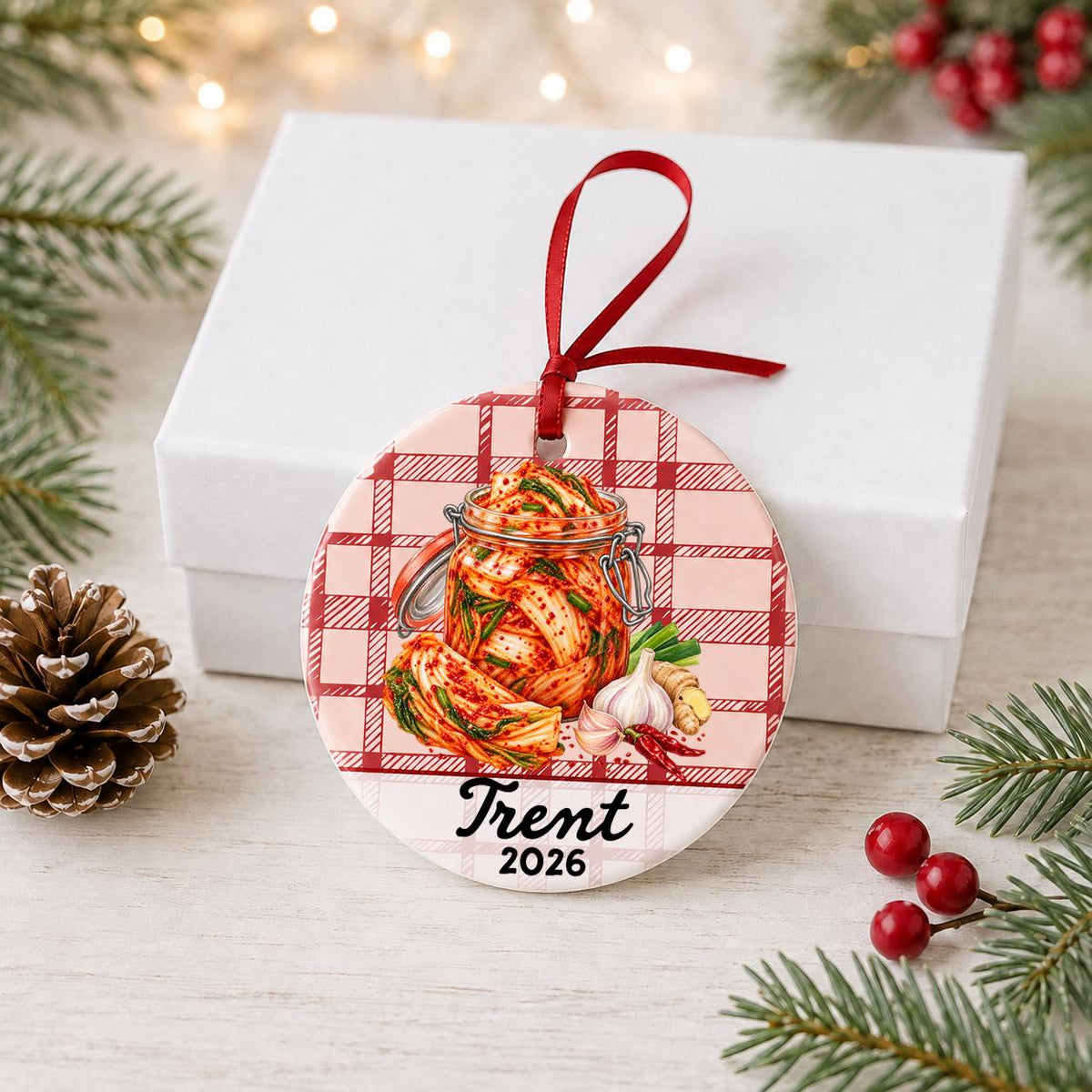 Personalized kimchi ceramic ornament with name and year on red plaid in front of a white gift box with pinecones and red berries