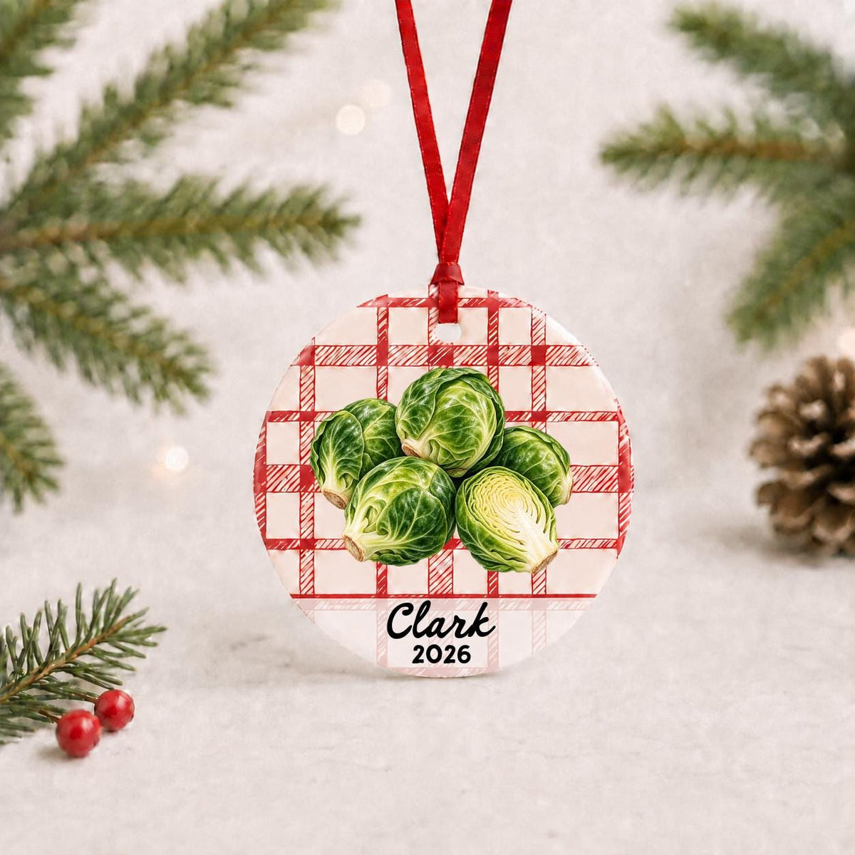 Personalized brussels sprouts ceramic ornament with name and year on red plaid with red ribbon on a white surface with pine branches and pinecones