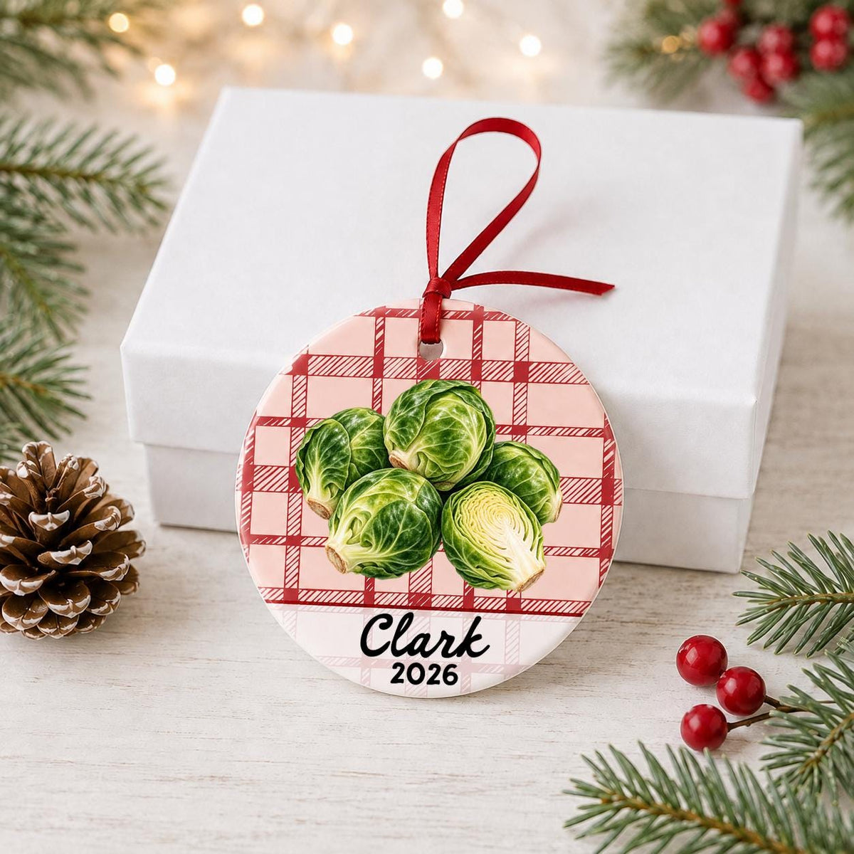 Personalized brussels sprouts ceramic ornament with name and year on red plaid in front of a white gift box with pinecones and red berries