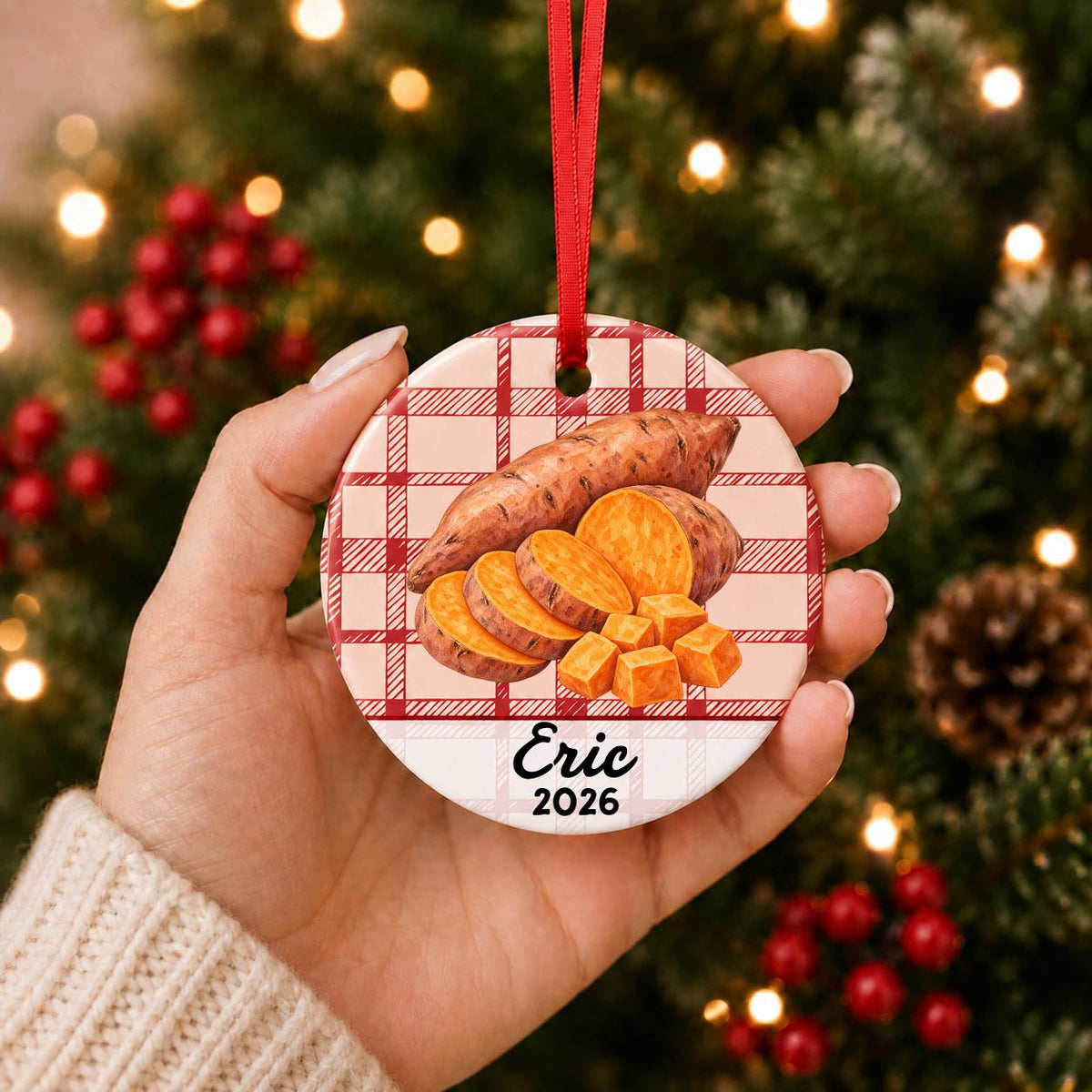 Hand holding the personalized sweet potato ceramic ornament with name and year on red plaid in front of a Christmas tree