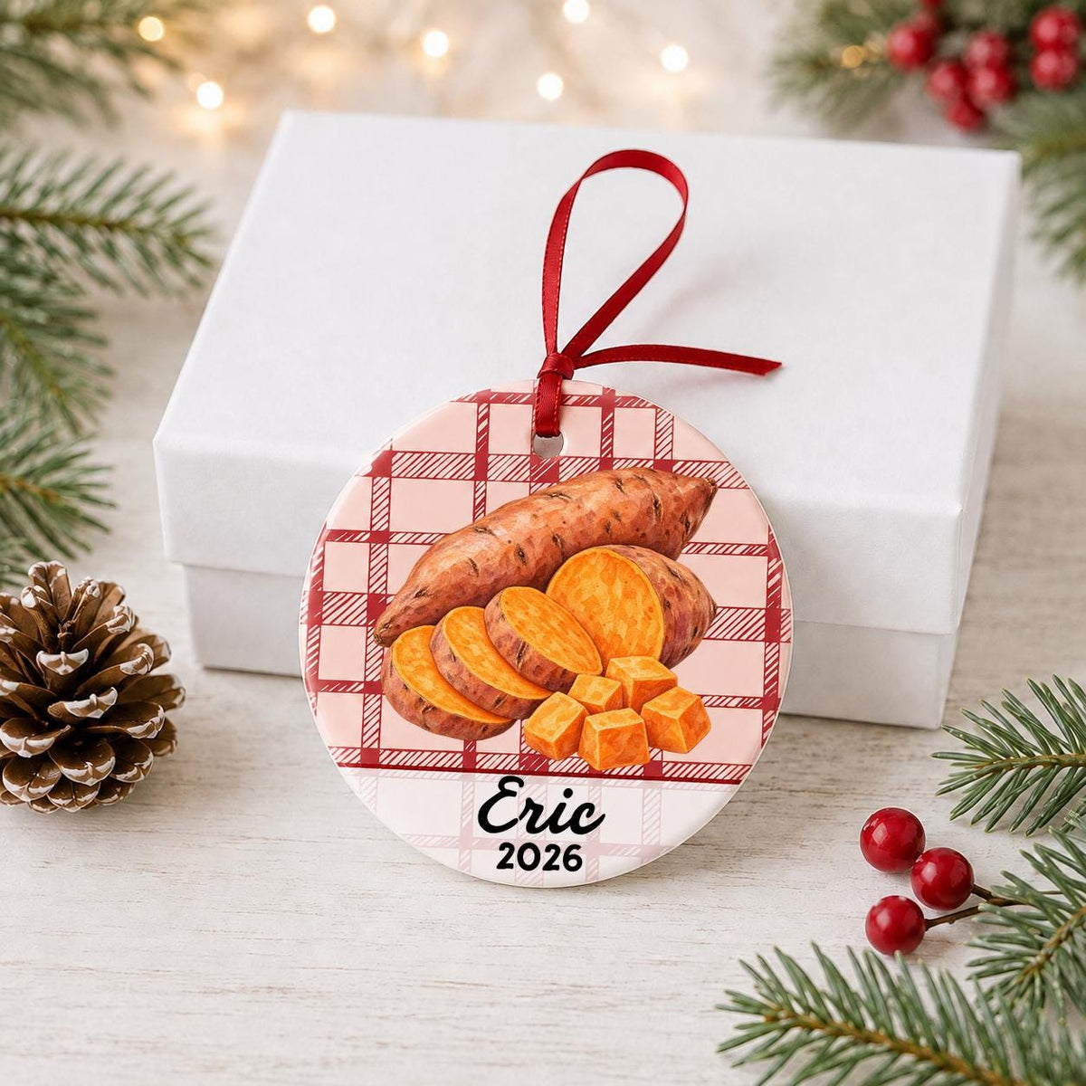 Personalized sweet potato ceramic ornament with name and year on red plaid in front of a white gift box with pinecones and red berries