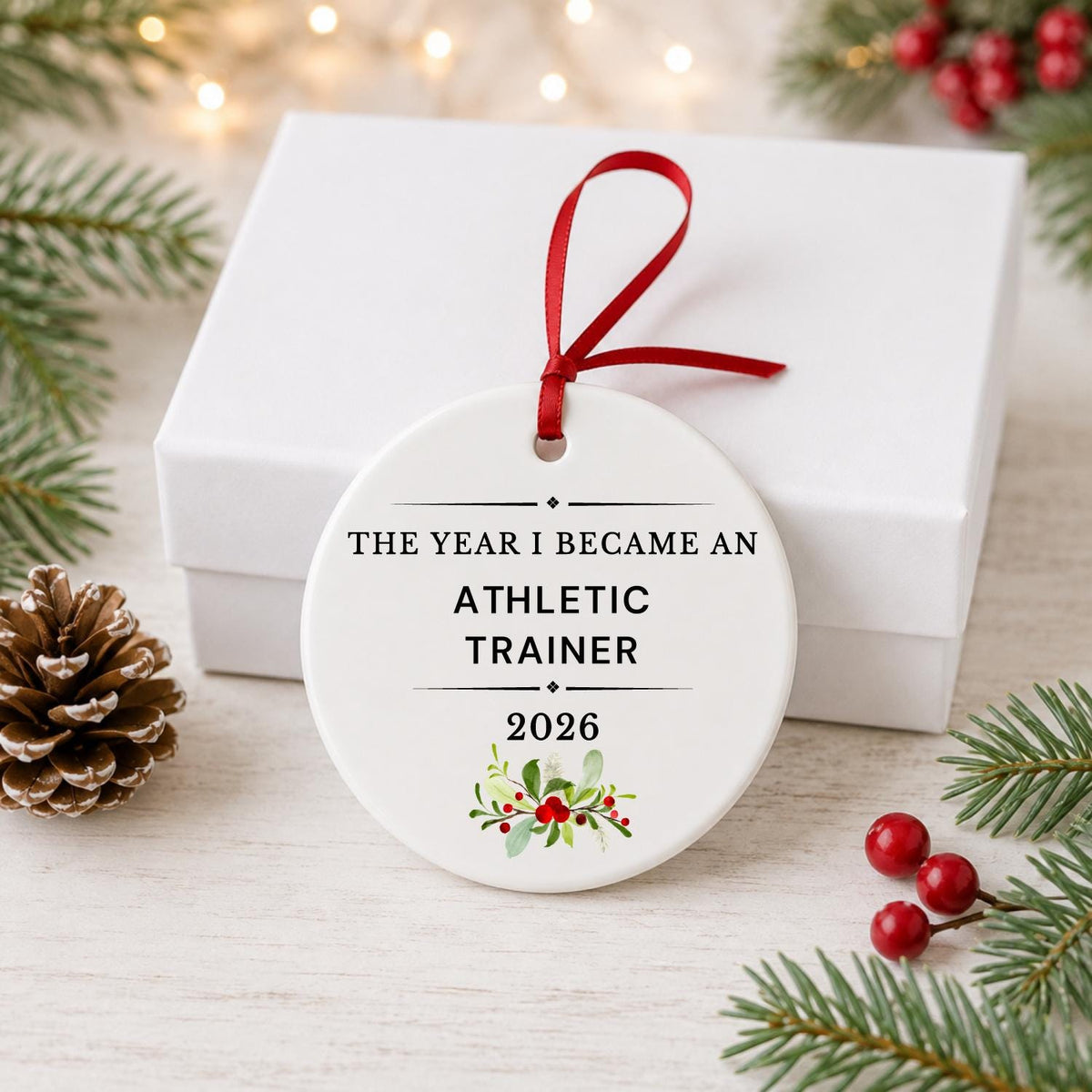 Athletic Trainer 2026 ceramic ornament reading The Year I Became an Athletic Trainer in front of a white gift box with pinecones and red berries