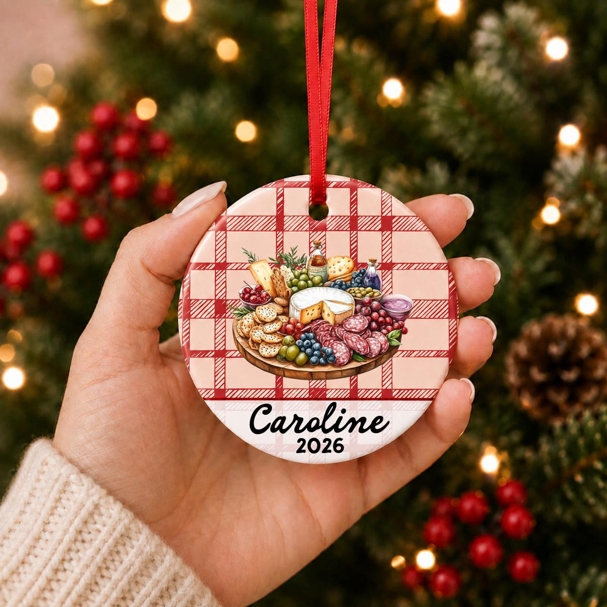 Hand holding the personalized charcuterie board ceramic ornament with name and year on red plaid in front of a Christmas tree