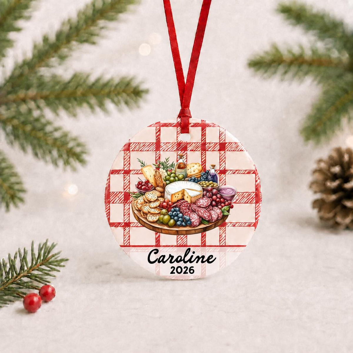 Personalized charcuterie board ceramic ornament with name and year on red plaid with red ribbon on a white surface with pine branches and pinecones
